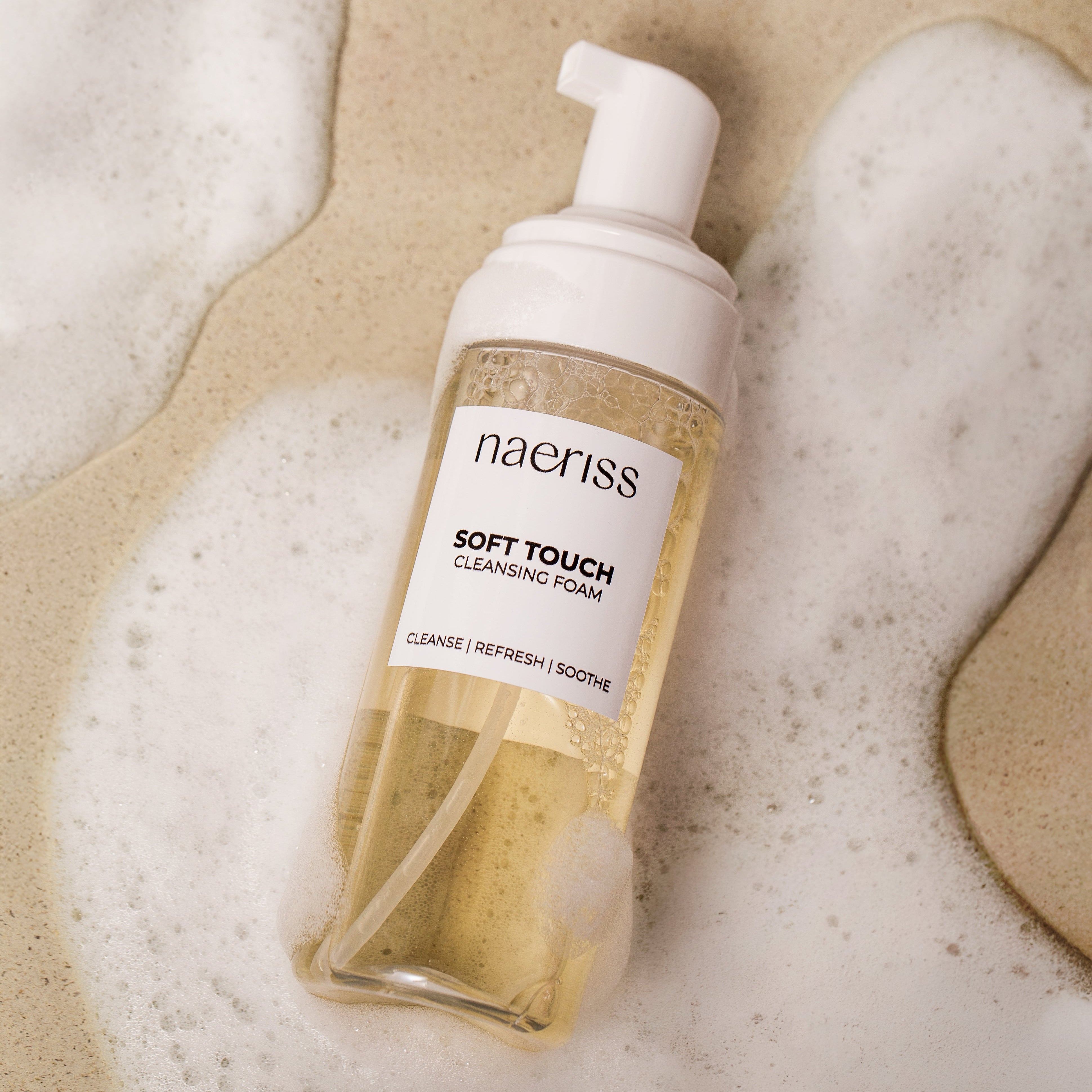 Naeriss Soft Touch Cleansing Foam bottle on a textured beige background with foam around it.